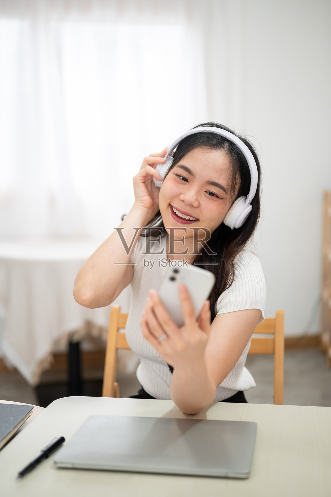 亚洲女性戴着耳机,坐在卧室的桌子旁,一边听音乐一边看手机。照片摄影图片