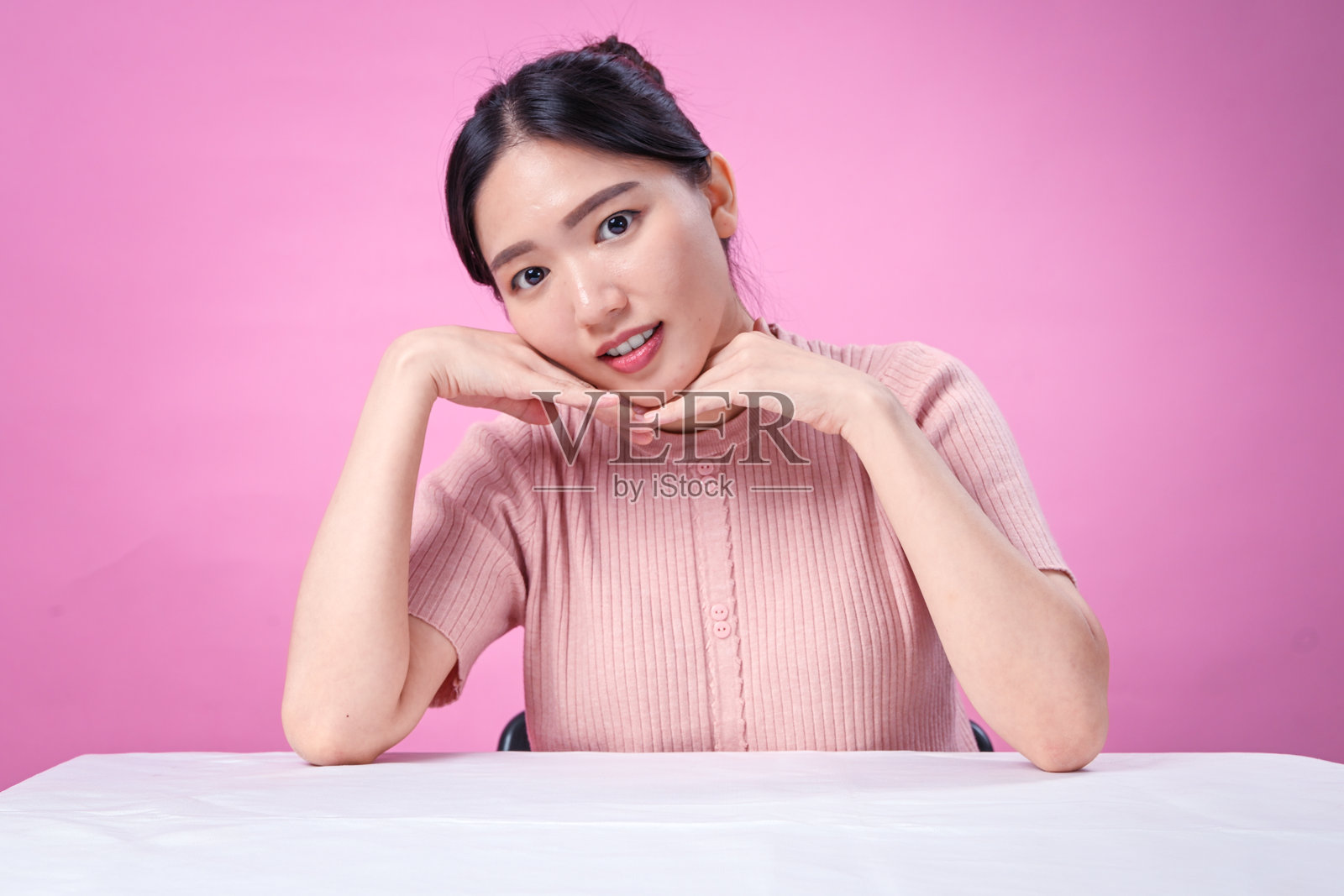 一位年轻的亚洲女性,留着深色头发,扎成马尾,身穿罗纹粉色短袖上衣,双手托腮,笑容灿烂,背景为纯粉色,表情显得十分愉悦。照片摄影图片