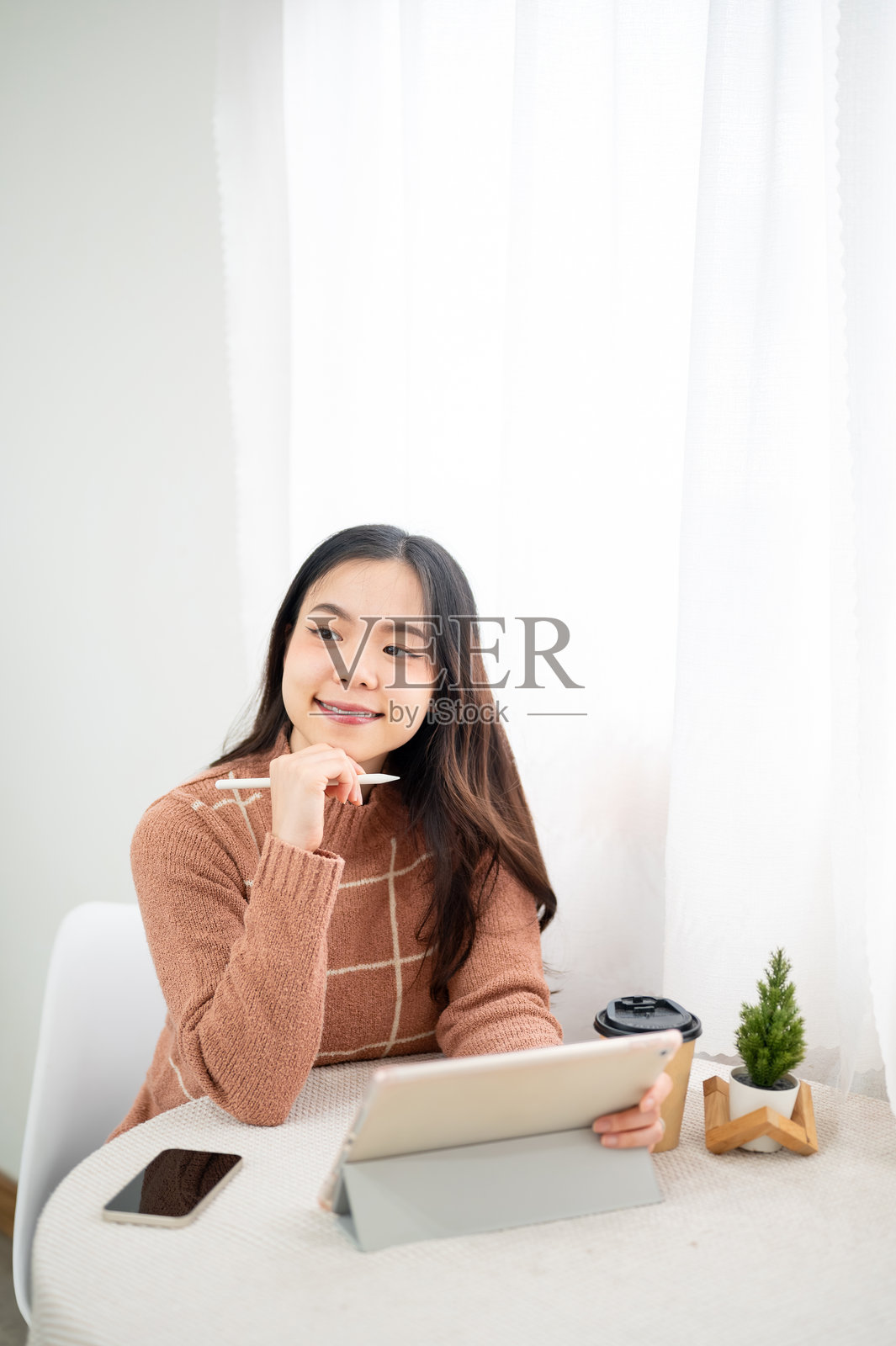 亚洲女性坐在白色房间的咖啡馆里,下巴托着触控笔,目光移开桌上的平板电脑。照片摄影图片