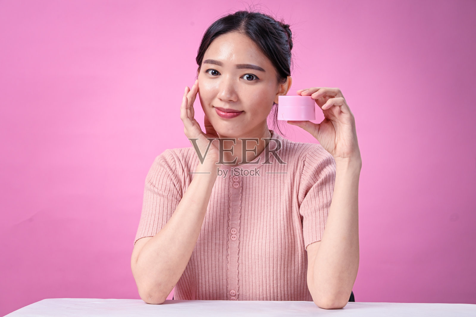 一位年轻的亚洲女性,梳着黑色发髻,穿着罗纹粉色短袖上衣,手持粉色护肤霜罐,轻触自己的脸庞,在柔和的粉色背景前推广护肤品照片摄影图片