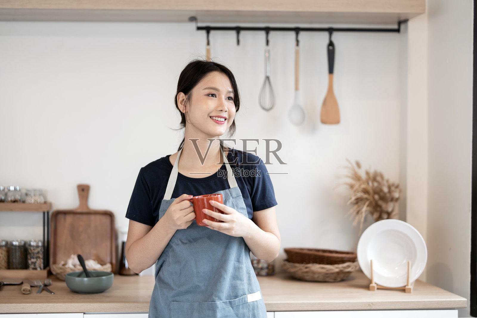 年轻的亚洲女性穿着围裙,在明亮现代的厨房里休息,享用热饮。照片摄影图片