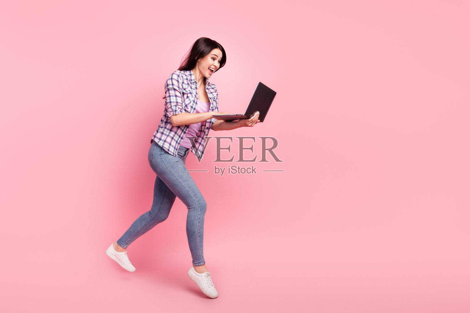 身穿格子衬衫的女孩在笔记本电脑上浏览网页,全身照,背景为隔离的粉彩色照片摄影图片