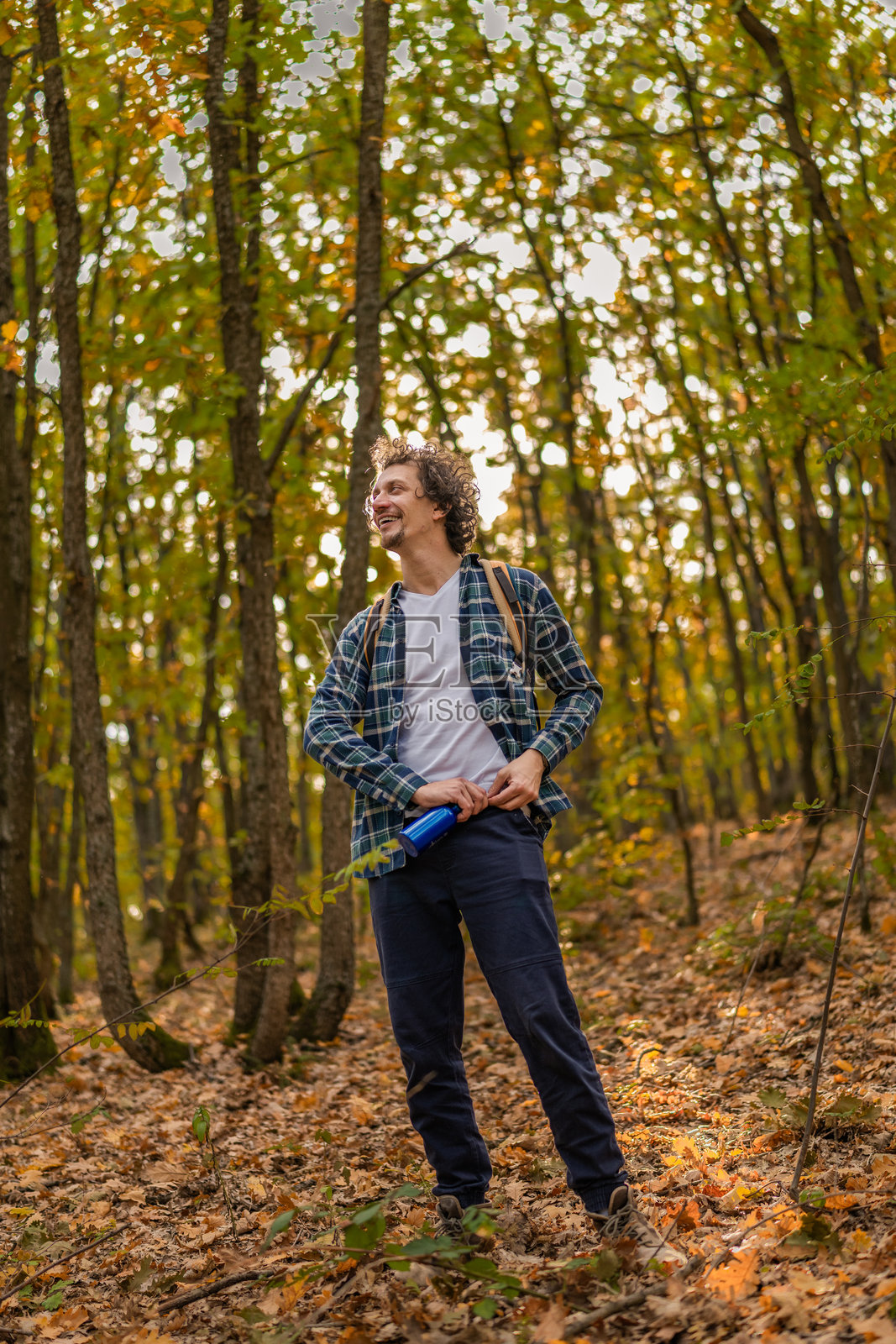 一个留着波浪卷发的男人走在森林里,打开一瓶水补充水分。照片摄影图片