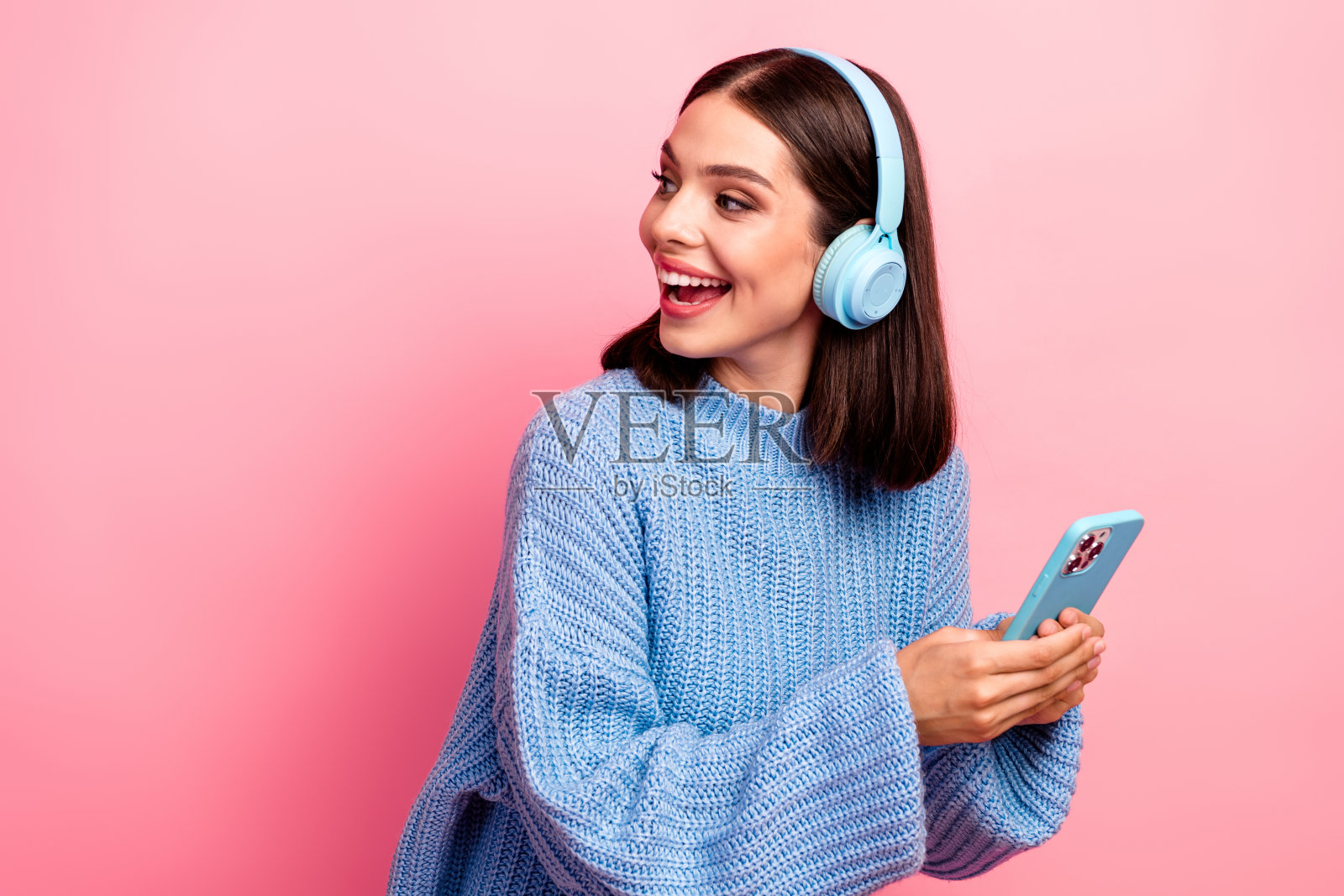 身穿蓝色毛衣的年轻女子戴着耳机,手持蓝色智能手机,背景为粉色照片摄影图片