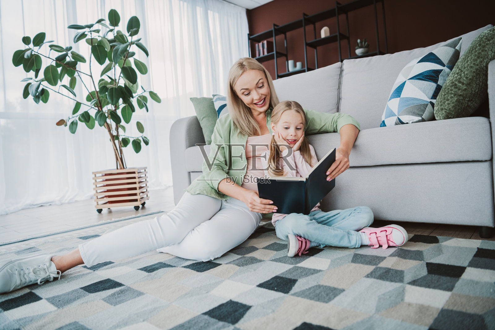 一位充满爱意的母亲和女儿在地板上一起阅读,舒适的沙发和绿色植物营造出温馨的家庭亲子时光,充满了学习的氛围。照片摄影图片