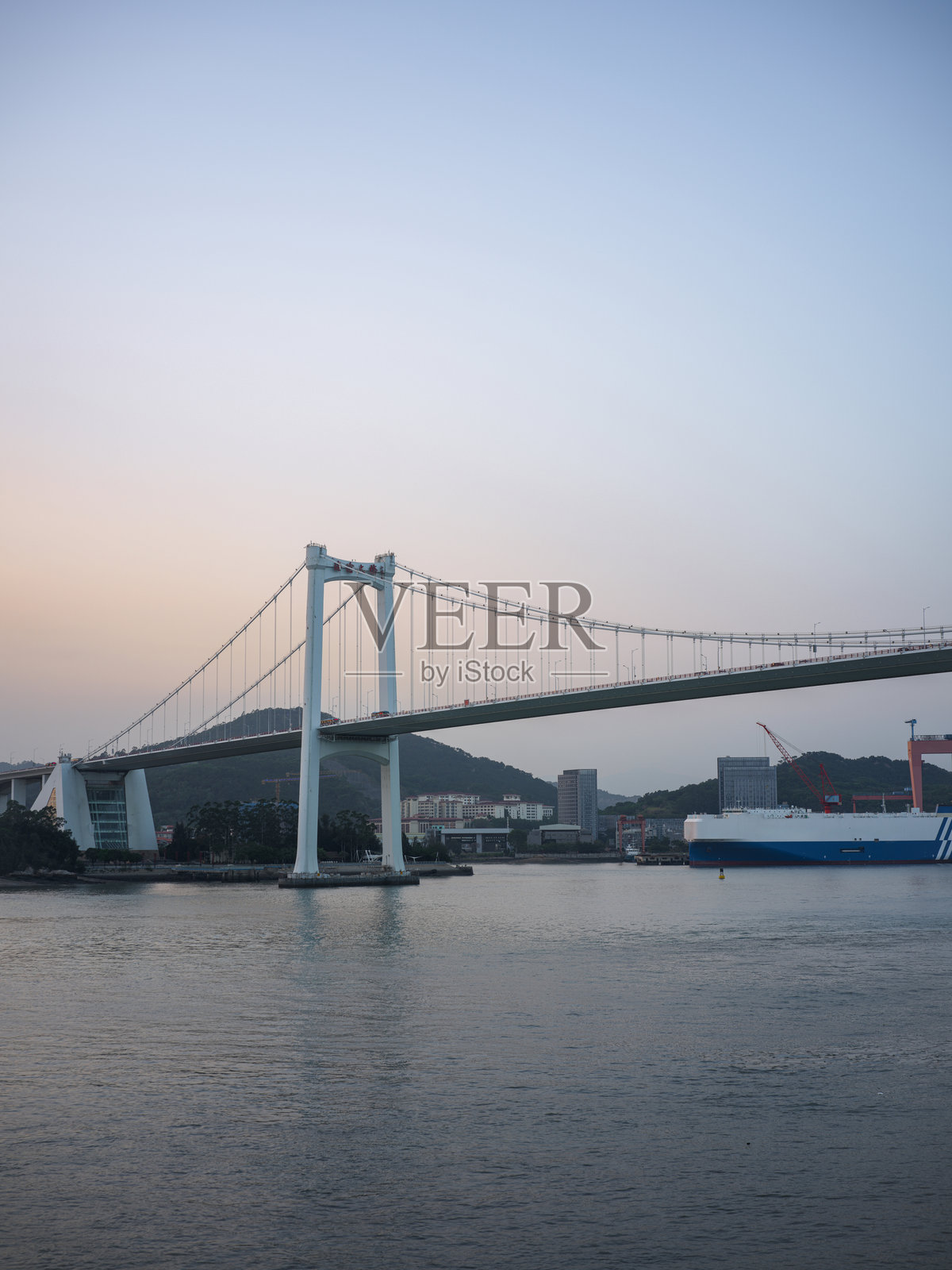 黄昏时分,一座壮观的悬索桥横跨大海,一艘货船停靠在港口。照片摄影图片