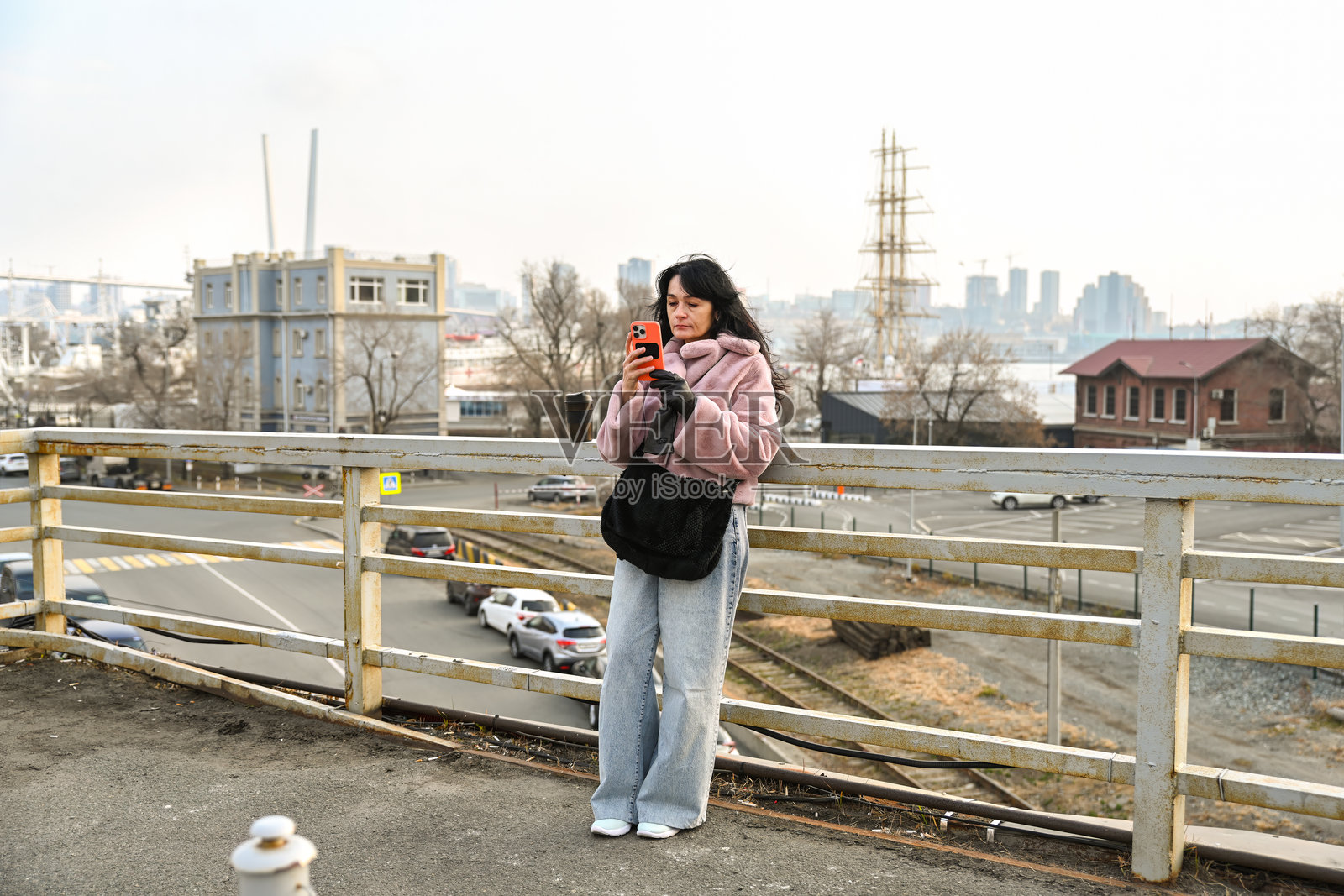 一位50岁的俄罗斯女性站在符拉迪沃斯托克的一个观景点,她正使用智能手机,在柔和的光线和城市景观中享受宁静的城市早晨。照片摄影图片