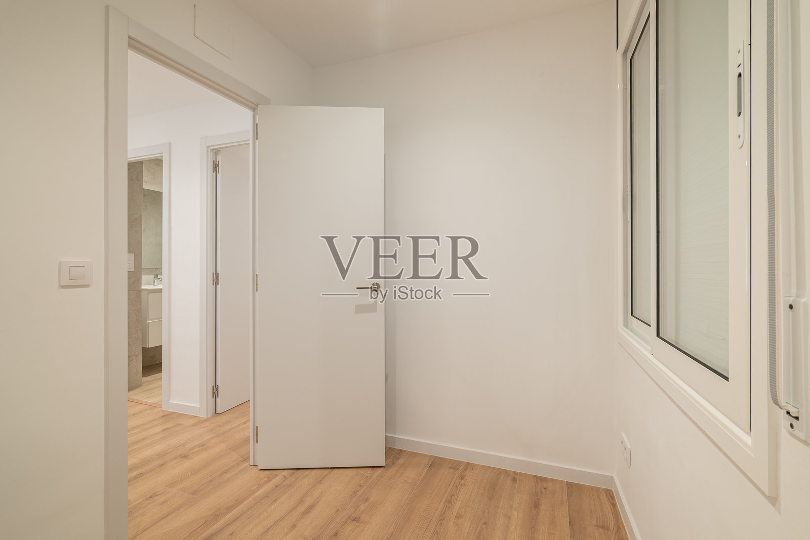 一个现代、空旷的房间,有白色的墙壁和木质地板。可以看到一扇关闭的白门,通向另一个空间。自然光从窗户射入。照片摄影图片