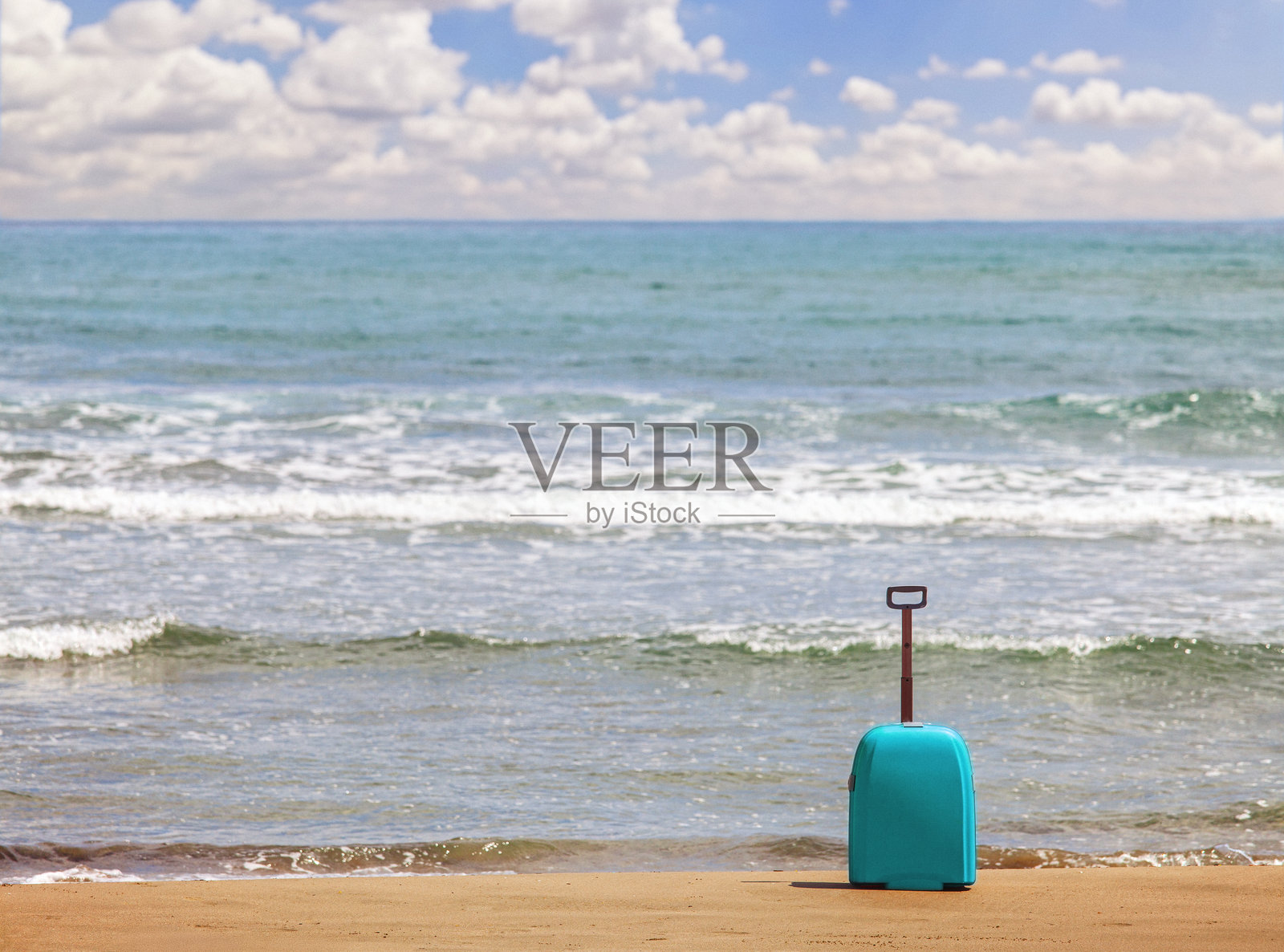 沙滩上的青绿色旅行箱,背景是轻柔的海浪、晴朗的蓝天和白云照片摄影图片