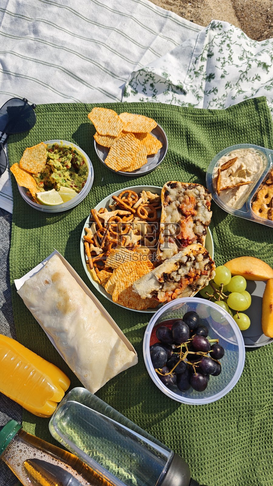 野餐用品,包括零食、水果、三明治和饮品,在夏日阳光下铺展在野餐垫上,画面传达出户外休闲、共享野餐和放松度假的愉悦感。照片摄影图片