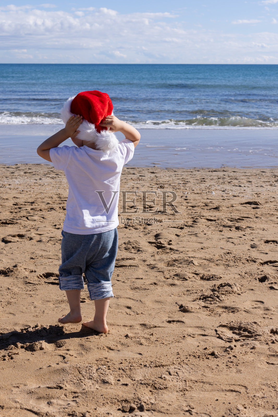 小男孩戴着红色的圣诞帽在海边行走。圣诞假期概念。照片摄影图片