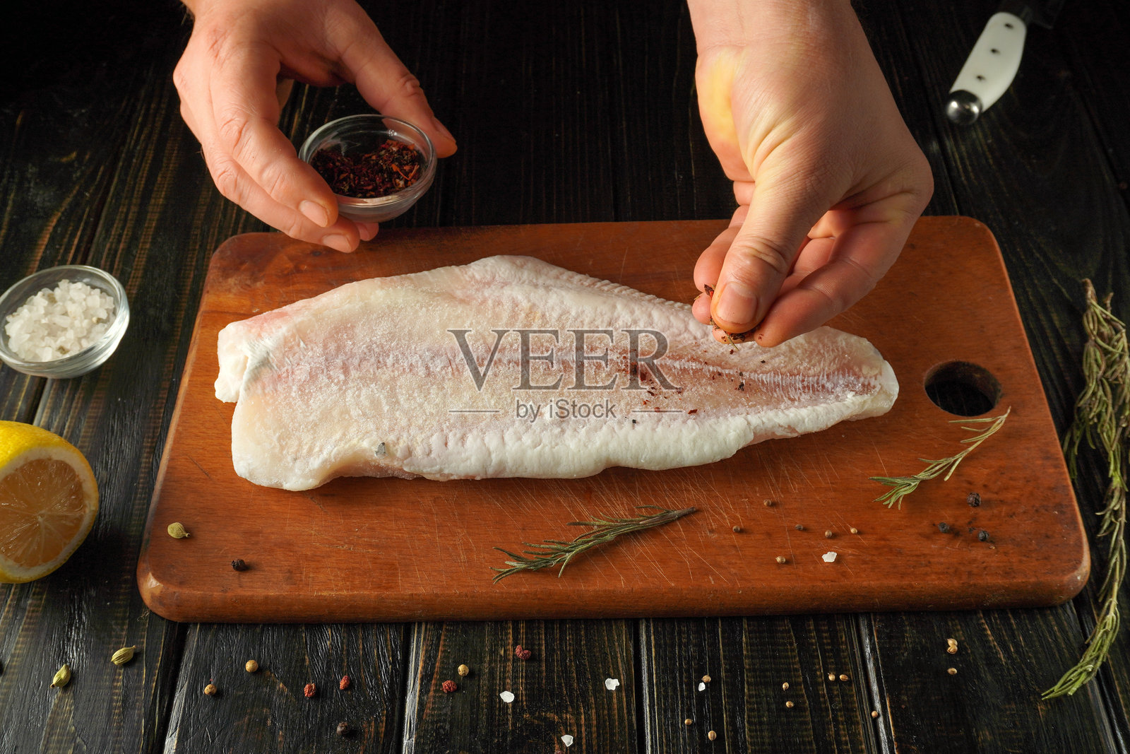 厨师正在仔细地用盐和香料调味新鲜的鱼片。柠檬和香草围绕在砧板旁,营造出备餐时色彩缤纷的厨房景象。照片摄影图片