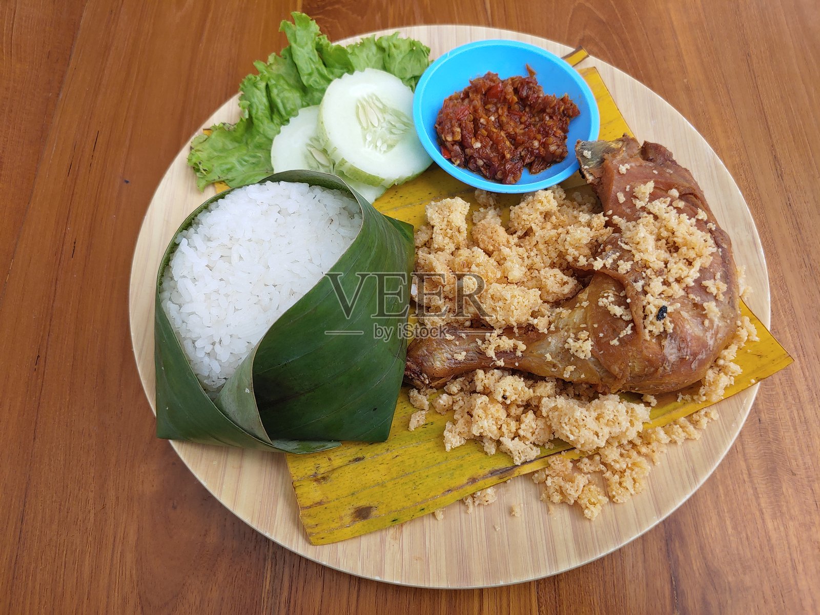 印尼菜:香脆炸鸡饭(Nasi Ayam Goreng Kremes),炸鸡用特制香料烹制,搭配酥脆的炸粉(kremes),用香蕉叶包裹米饭,配以黄瓜片、生菜和酱汁照片摄影图片