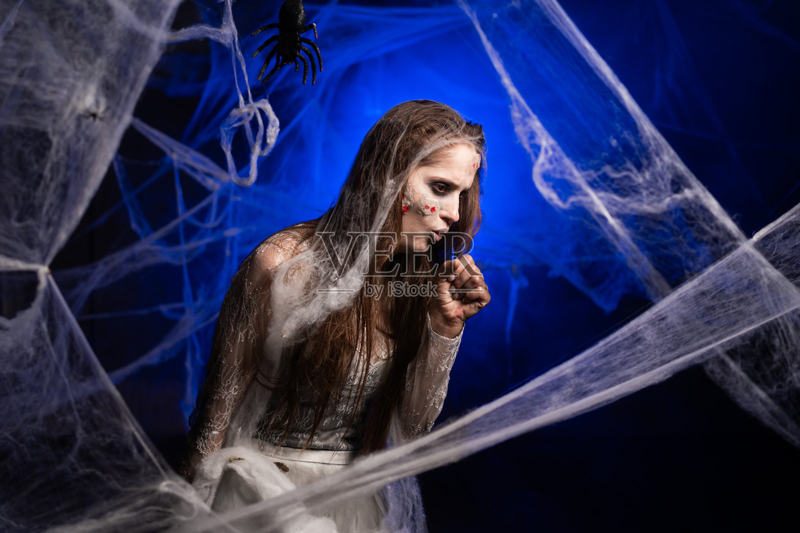 治疗,医疗保健概念。生病的邪恶女子,化着恐怖吓人的万圣节僵尸妆,正在咳嗽,感染了病毒或感冒,站在蓝色背景前照片摄影图片