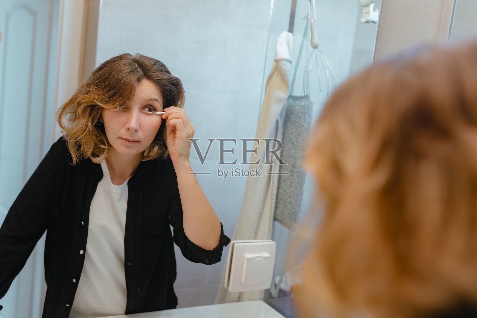 女性在镜子前描画眼线,浴室暖光,日常美容,专注的表情,自然妆容,柔顺的棕色头发,休闲的着装。照片摄影图片