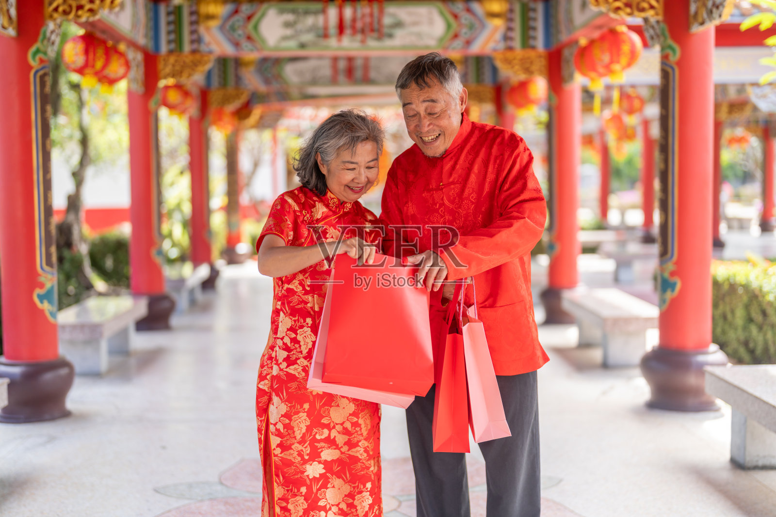春节期间,一对快乐的亚洲老年夫妇在中国寺庙里拿着红色的购物袋,年迈的丈夫和妻子穿着旗袍笑着庆祝购物节。退休生活方式的概念照片摄影图片