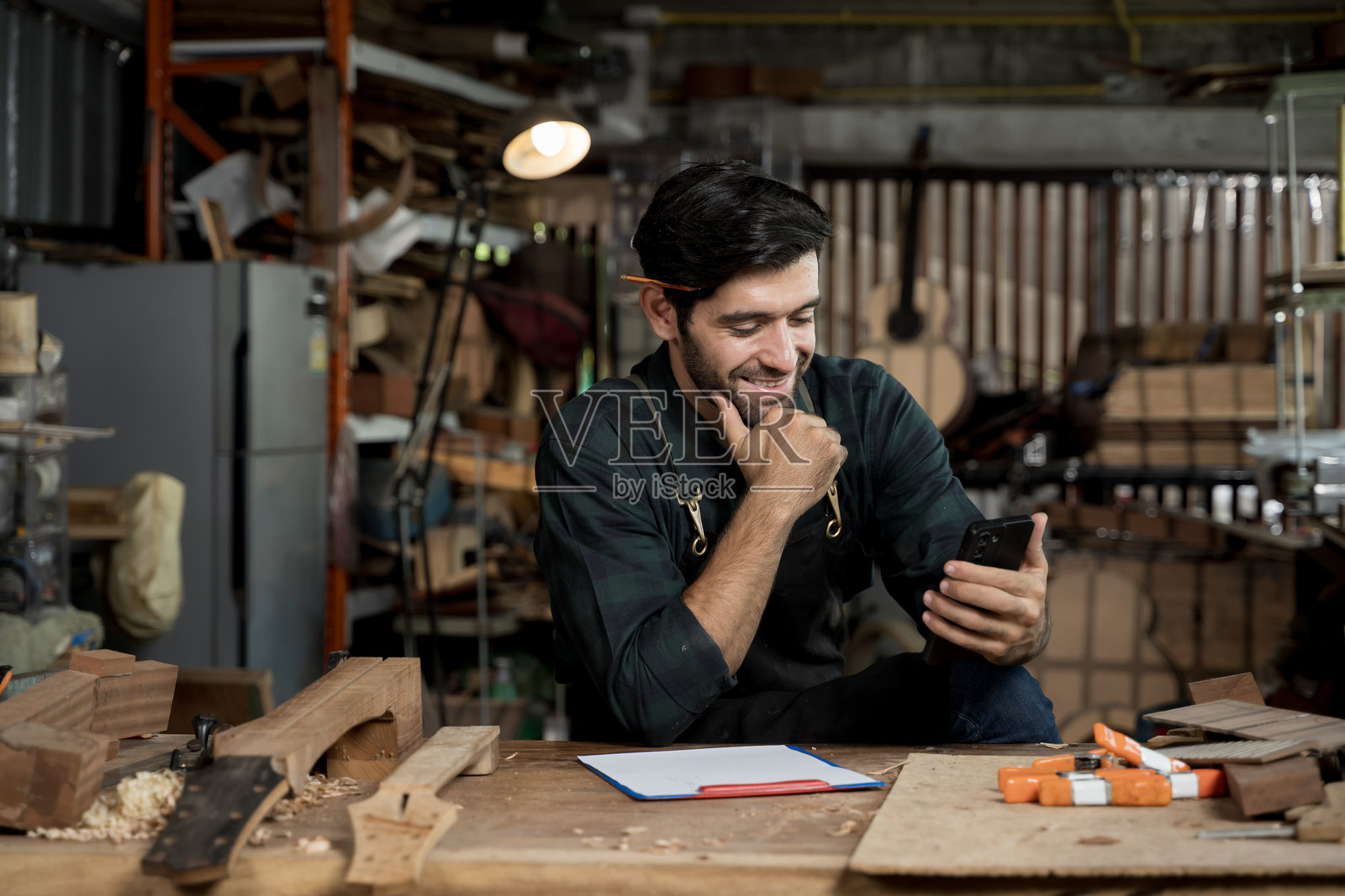 工作坊里,一位面带微笑的鲁特琴制造师正在看手机上的消息,一位小企业工匠在他的工作台前。照片摄影图片