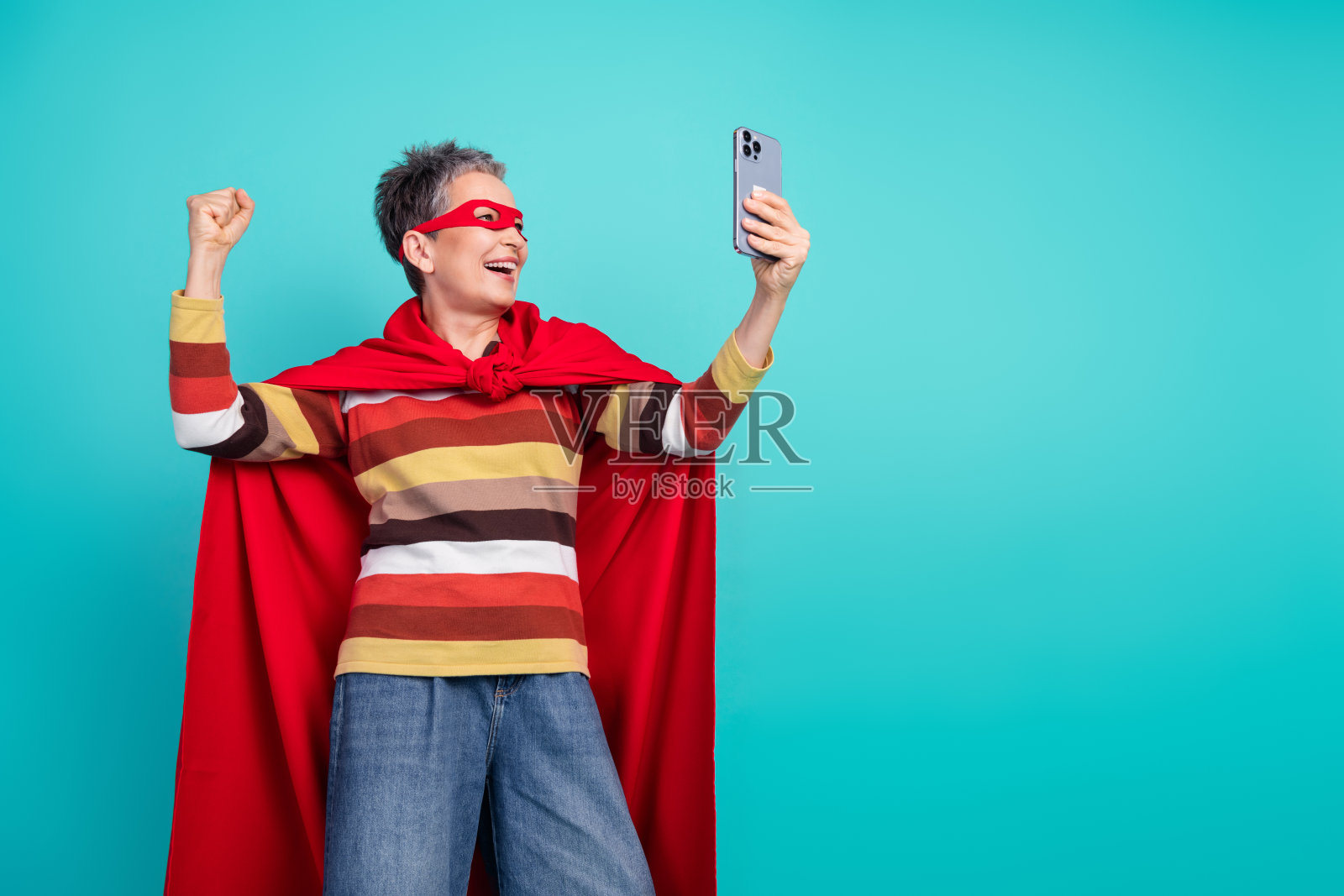 一位身穿条纹毛衣、披着红色斗篷的成熟的女性,以超级英雄的风格在蓝色背景前自拍。照片摄影图片