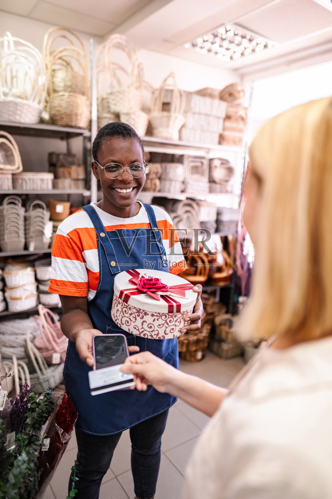 微笑的女销售员手持礼品盒,接受顾客使用智能手机和信用卡付款照片摄影图片