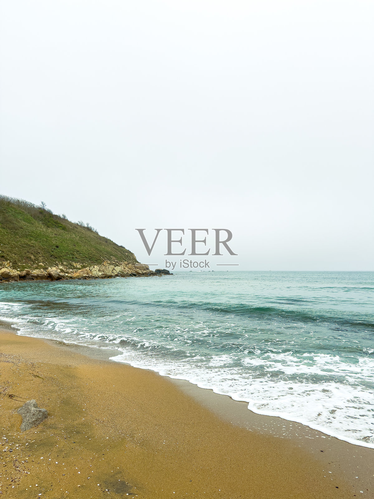阴天时,海滩边绿色的山坡旁,有沙滩和海浪拍打的海岸线照片摄影图片
