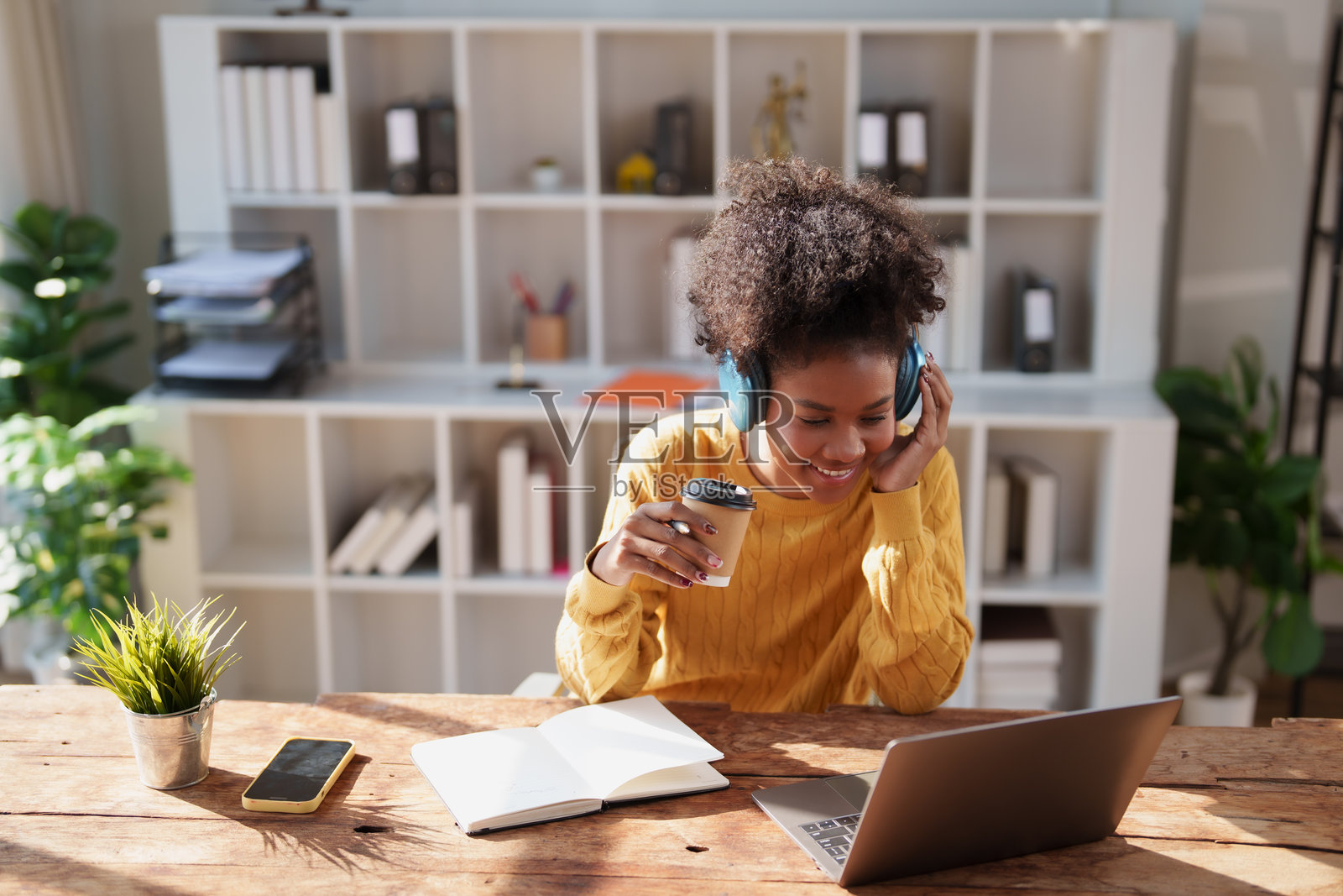 年轻女性在家中办公室学习、远程工作或参加在线会议,她正专注于笔记本电脑,同时享用咖啡并戴着耳机听讲。照片摄影图片