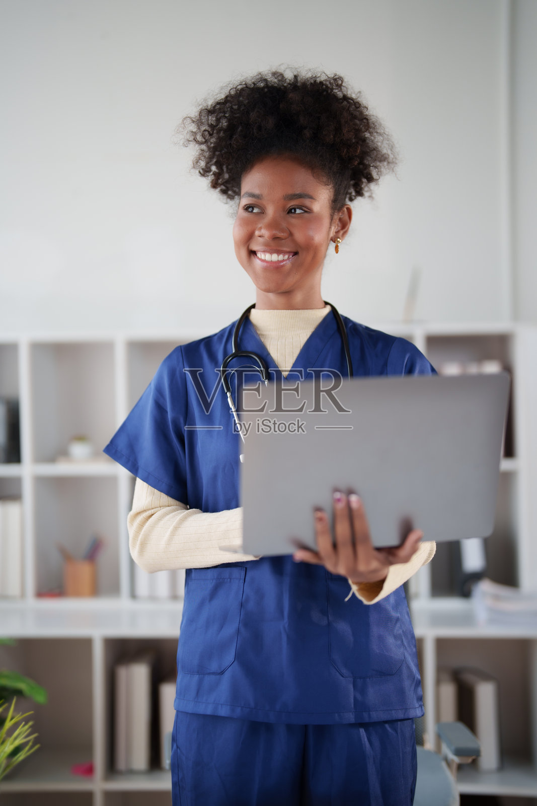 一位年轻的非裔美国护士从业者,身穿工作服,戴着听诊器,在现代医疗诊所自信地拿着笔记本电脑,脸上带着微笑。照片摄影图片