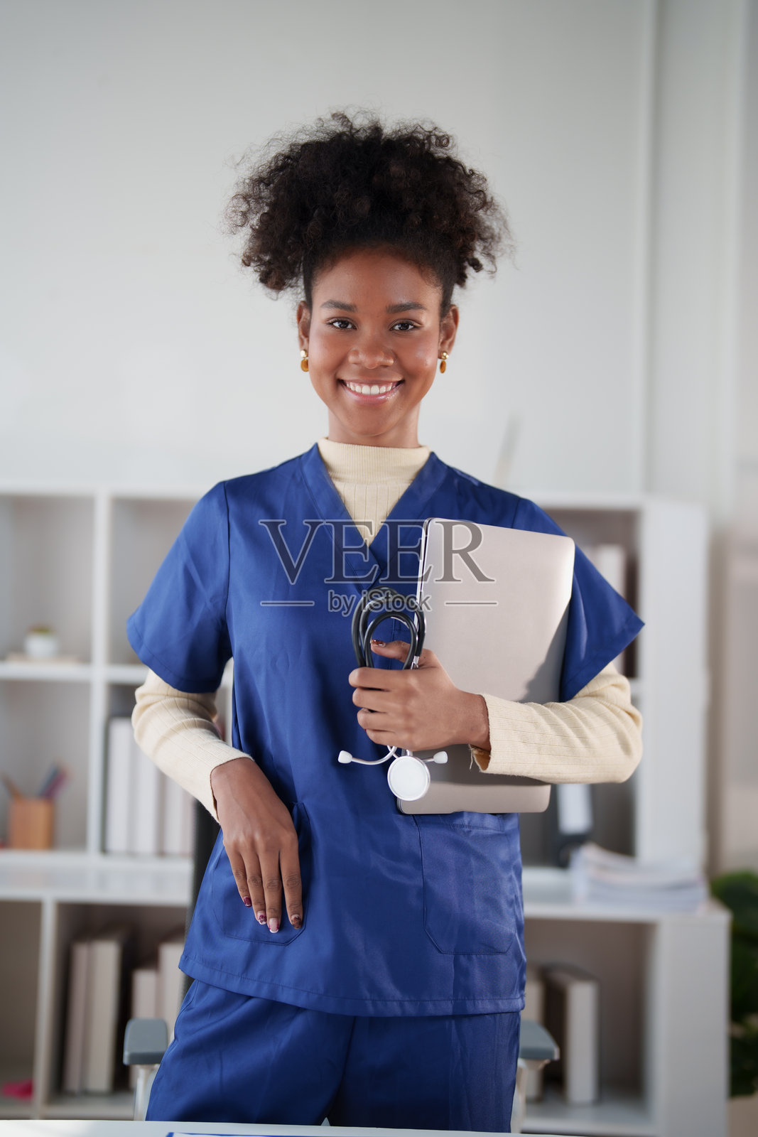 一位年轻的非裔美国护士自信地穿着工作服,手持笔记本电脑和听诊器,代表着医疗保健、科技和职业成功。照片摄影图片