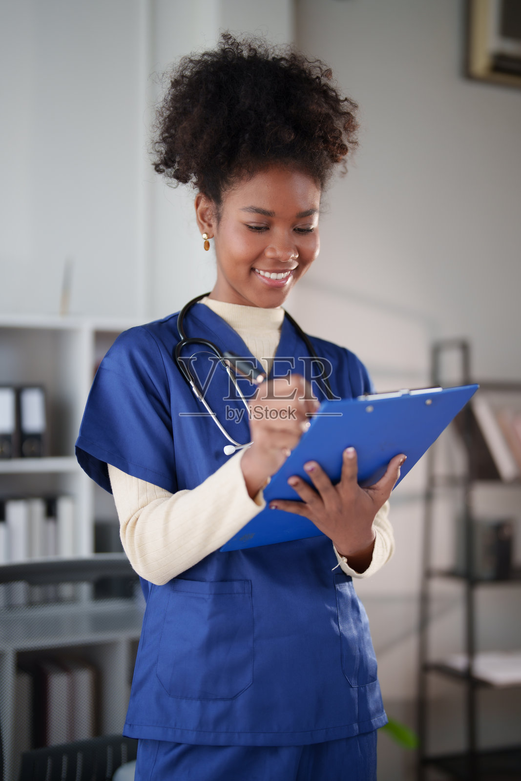 一位面带微笑的年轻非洲裔美国护士,身穿手术服,戴着听诊器,正用笔在蓝色的文件夹上书写,在医院或诊所环境中查阅病人病历。照片摄影图片