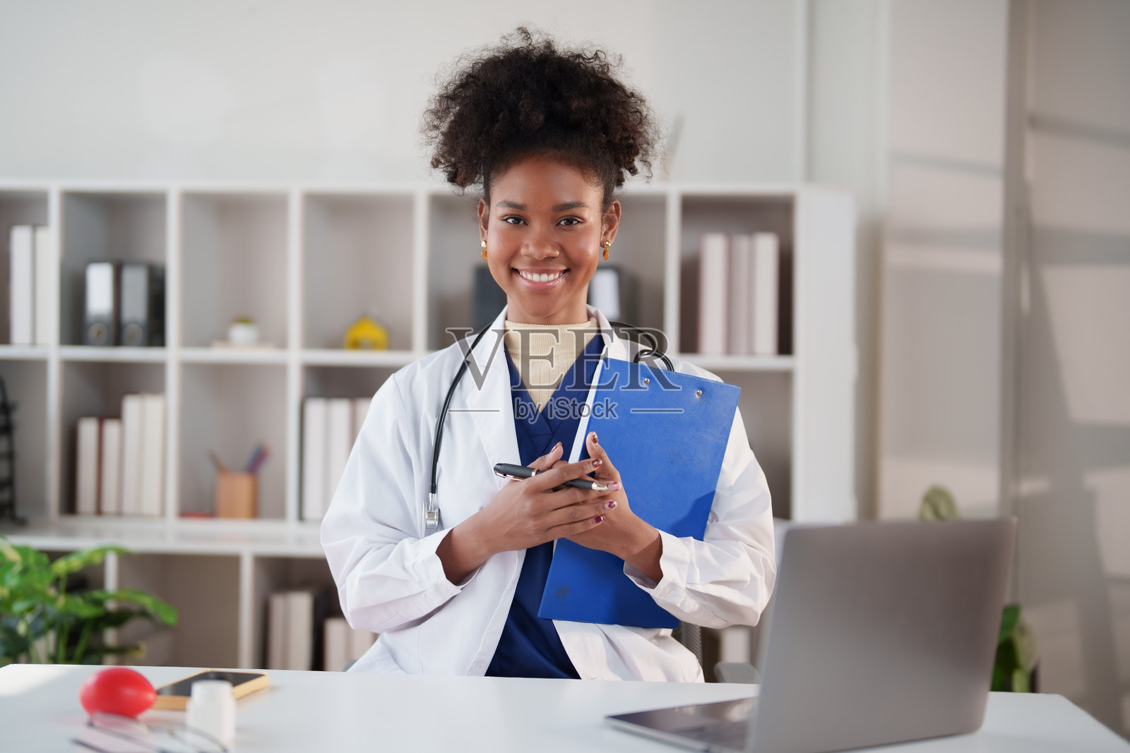 一位年轻的女医生,身穿实验服,戴着听诊器,手持文件夹和笔,自信地对着镜头微笑,身处一家现代诊所,传递出专业关怀和专业知识。照片摄影图片