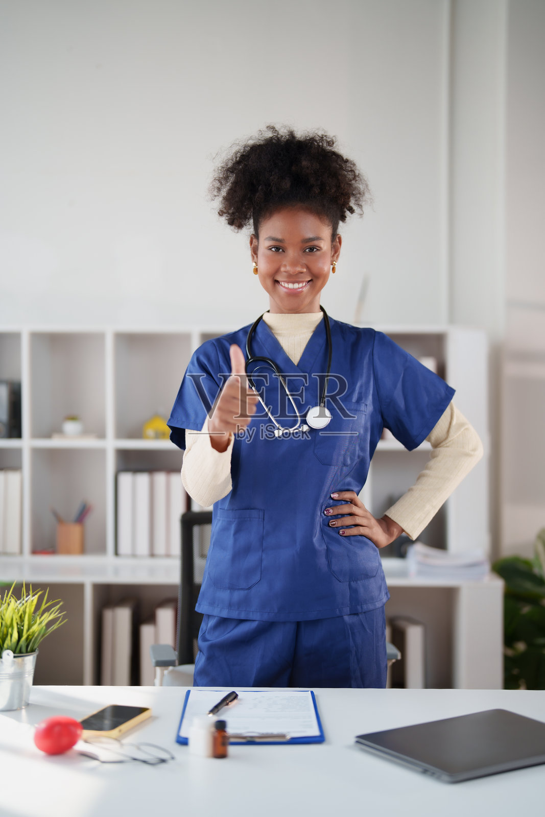 一位年轻的非洲裔女性医疗专业人士,身穿蓝色手术服,在诊所办公室里微笑着竖起大拇指,她自信且平易近人,身后是听诊器和办公桌。照片摄影图片