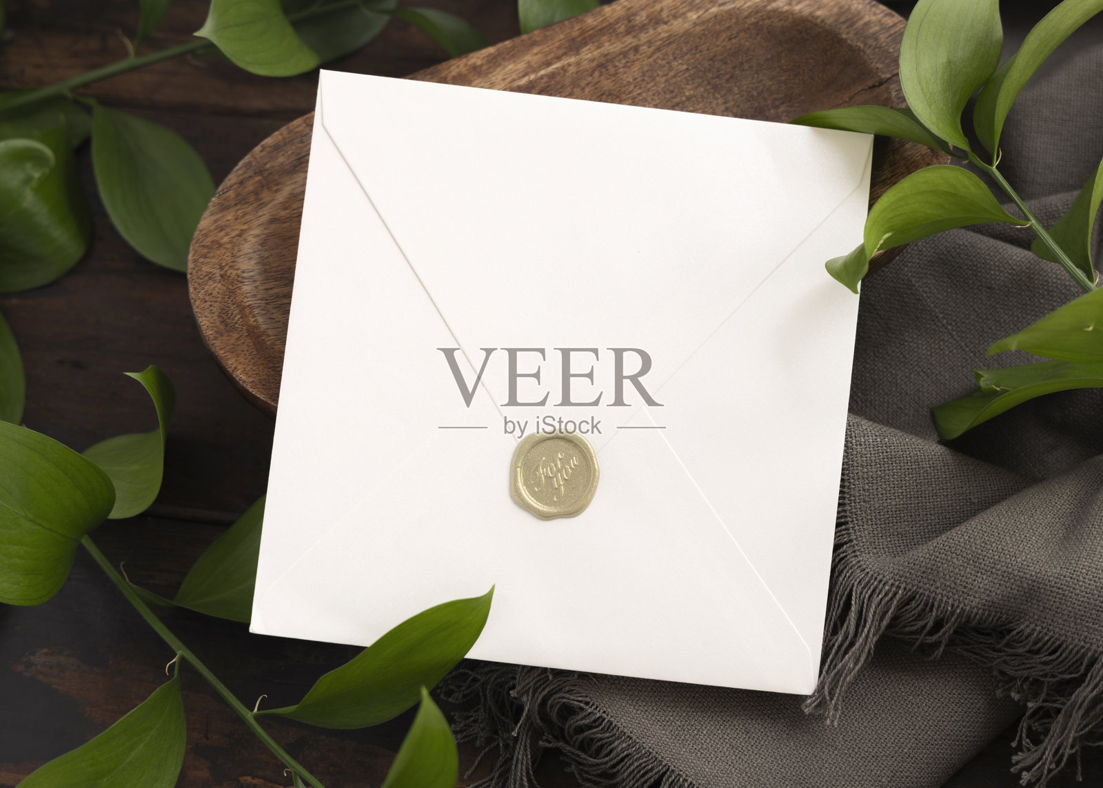密封的信封模型放在棕色木托盘上,旁边是绿叶和灰色织物,俯视角度照片摄影图片