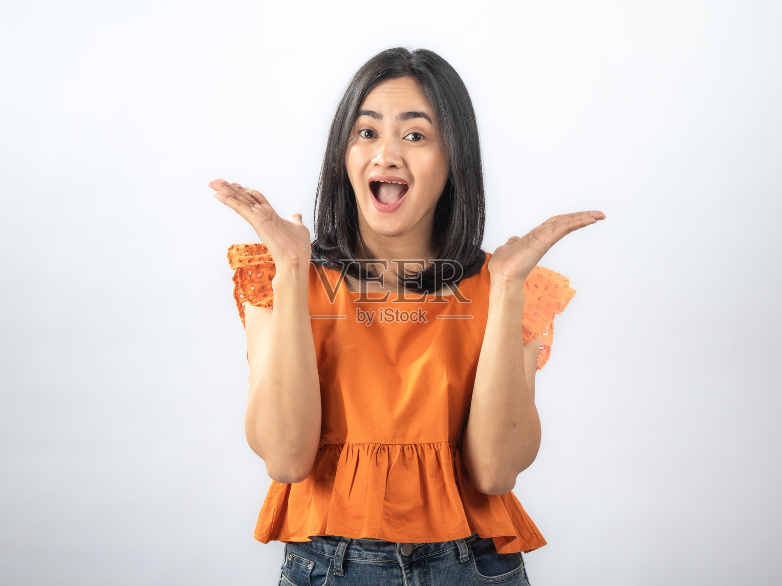 一位留着黑色直发的东南亚女性,身穿橙色荷叶边衬衫和牛仔裤,面带喜悦的表情,双手举在脸颊两侧,背景为纯白色。照片摄影图片