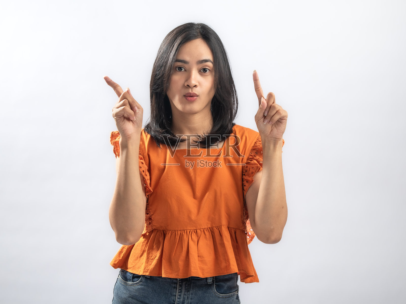 一位东南亚女性,留着黑色直发,身穿橙色荷叶边衬衫和牛仔裤,表情严肃,双手食指向上举起,背景为纯白色照片摄影图片