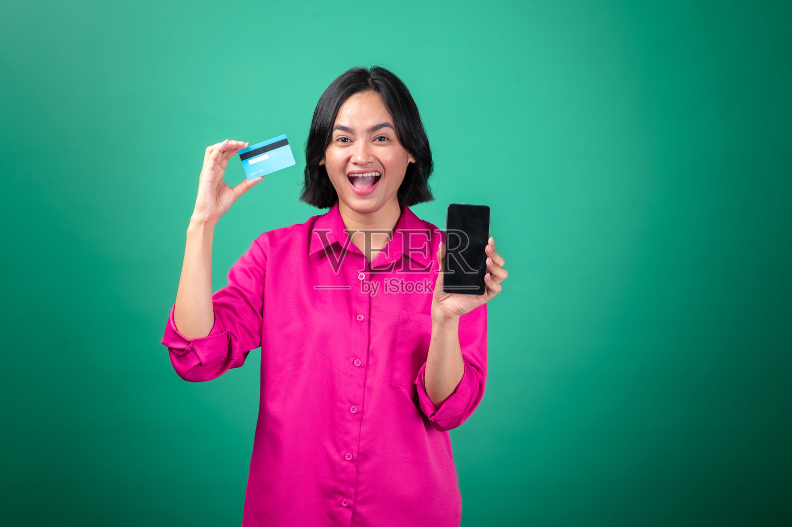 一位穿着粉色衬衫的亚洲女性,背景为绿色,她一只手拿着信用卡,另一只手拿着智能手机。她热情地微笑着,象征着在线购物或数字支付。照片摄影图片