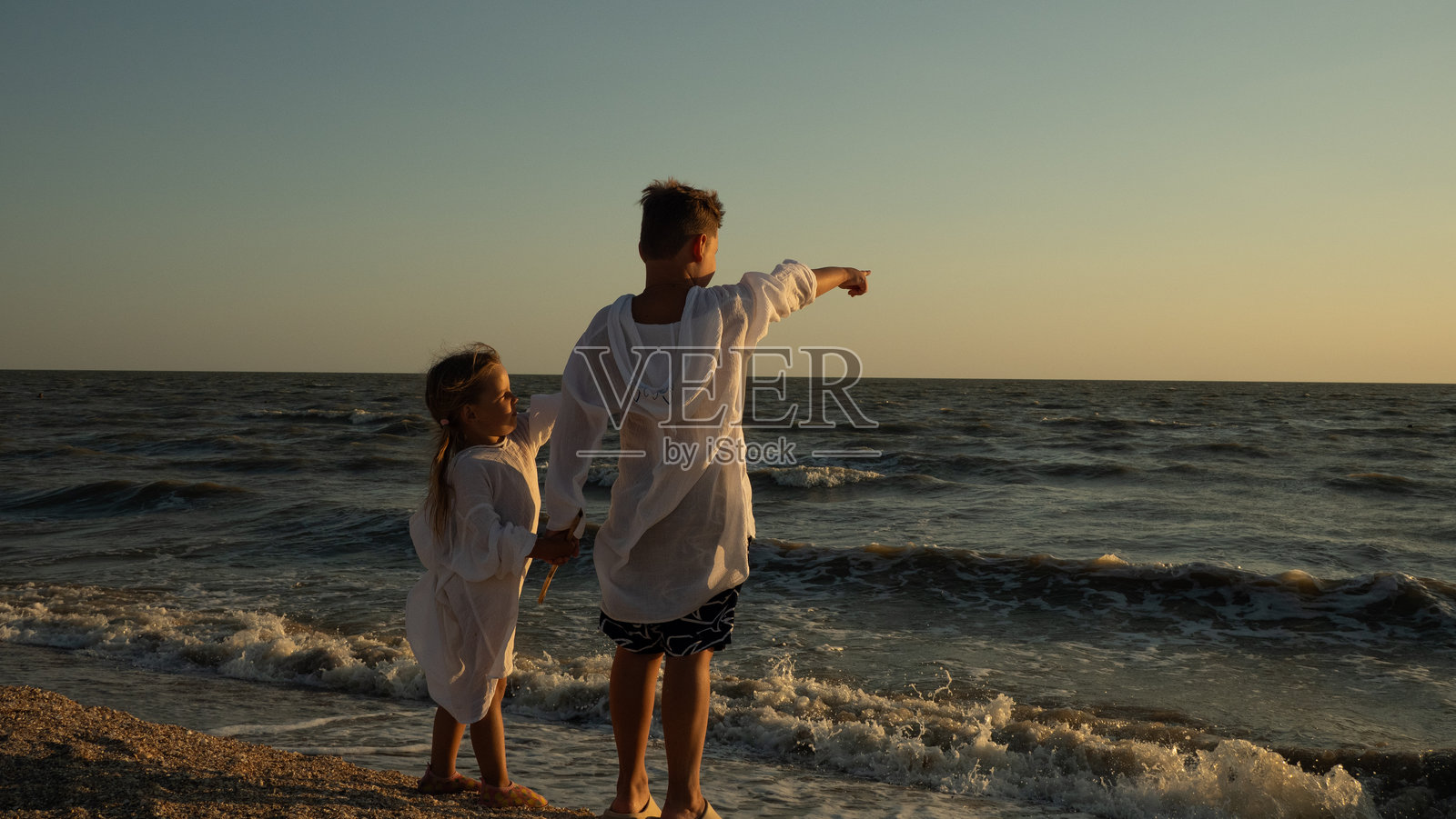一对姐弟(兄妹)在日落时分并肩站立在沙滩上,手牵着手,指向辽阔的大海。孩子们在海边共享着一份对自然的赞美和与自然的连接。照片摄影图片