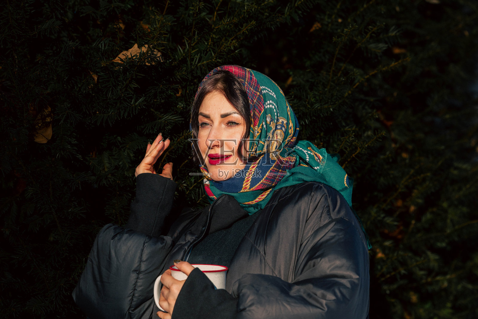 一位深色头发的女士,围着彩色围巾,穿着宽大的外套,手里拿着杯子,站在户外常绿的树丛中,在宁静的氛围中散发出温暖和舒适感。照片摄影图片