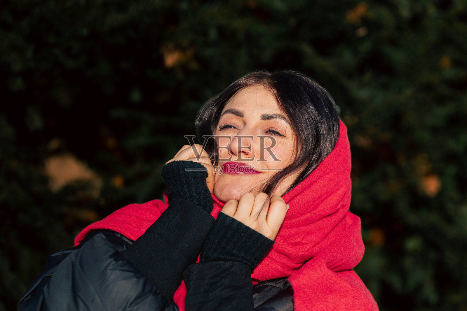 一位黑发年轻女子,戴着红色围巾,身穿黑色夹克,在户外开怀微笑,周围绿意盎然,捕捉了自然环境中幸福与温暖的精髓照片摄影图片