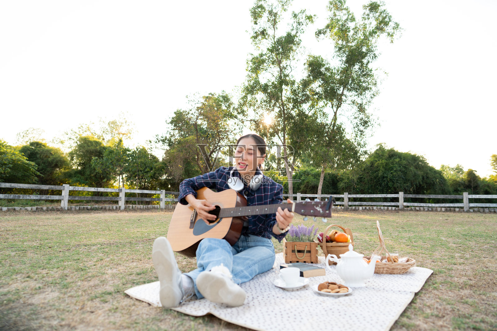 日落时分,快乐的亚洲女性在公园的野餐毯上弹奏原声吉他放松,体现生活方式和音乐爱好理念照片摄影图片
