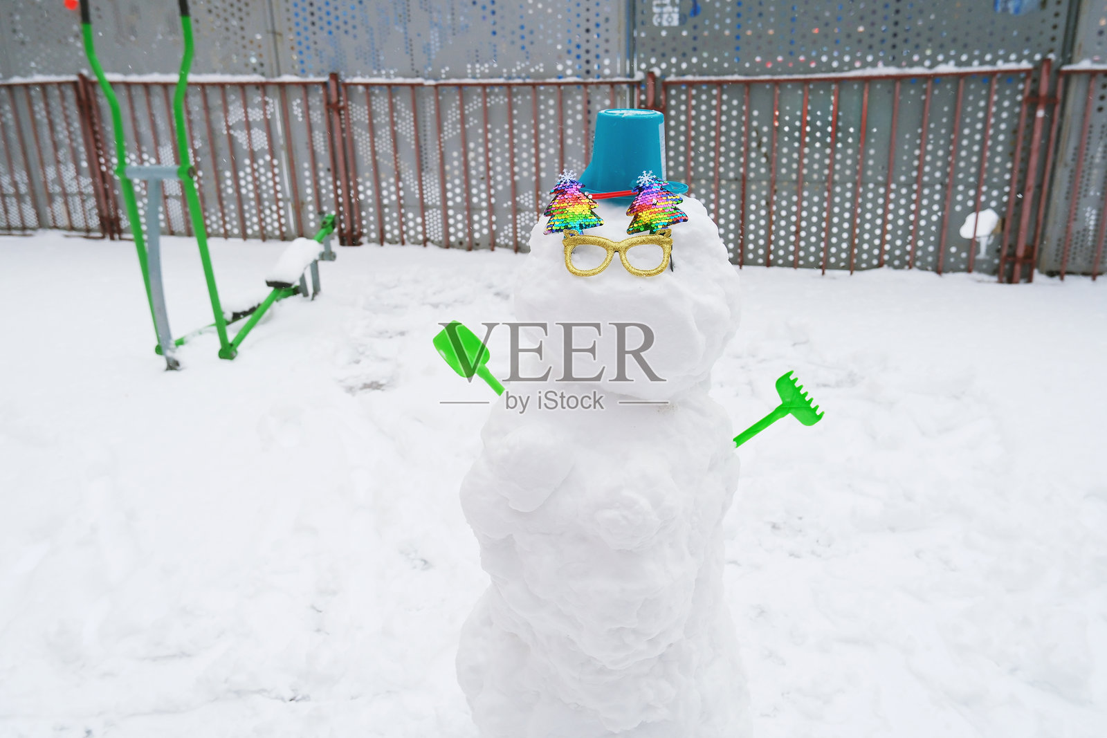 一个异想天开的雪人,装饰着多彩的饰品和一顶桶帽,在雪地游乐场里拿着绿色工具。一副亮色的太阳镜为这冬季景象增添了趣味。照片摄影图片