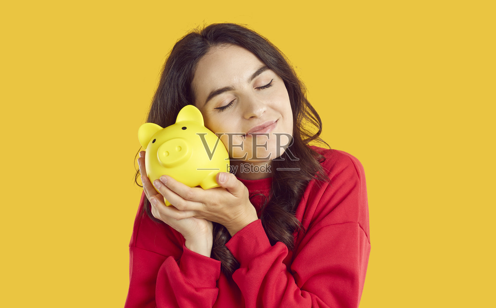 微笑的年轻女子轻轻地将脸颊贴近存钱罐,背景为橙色,孤立。照片摄影图片