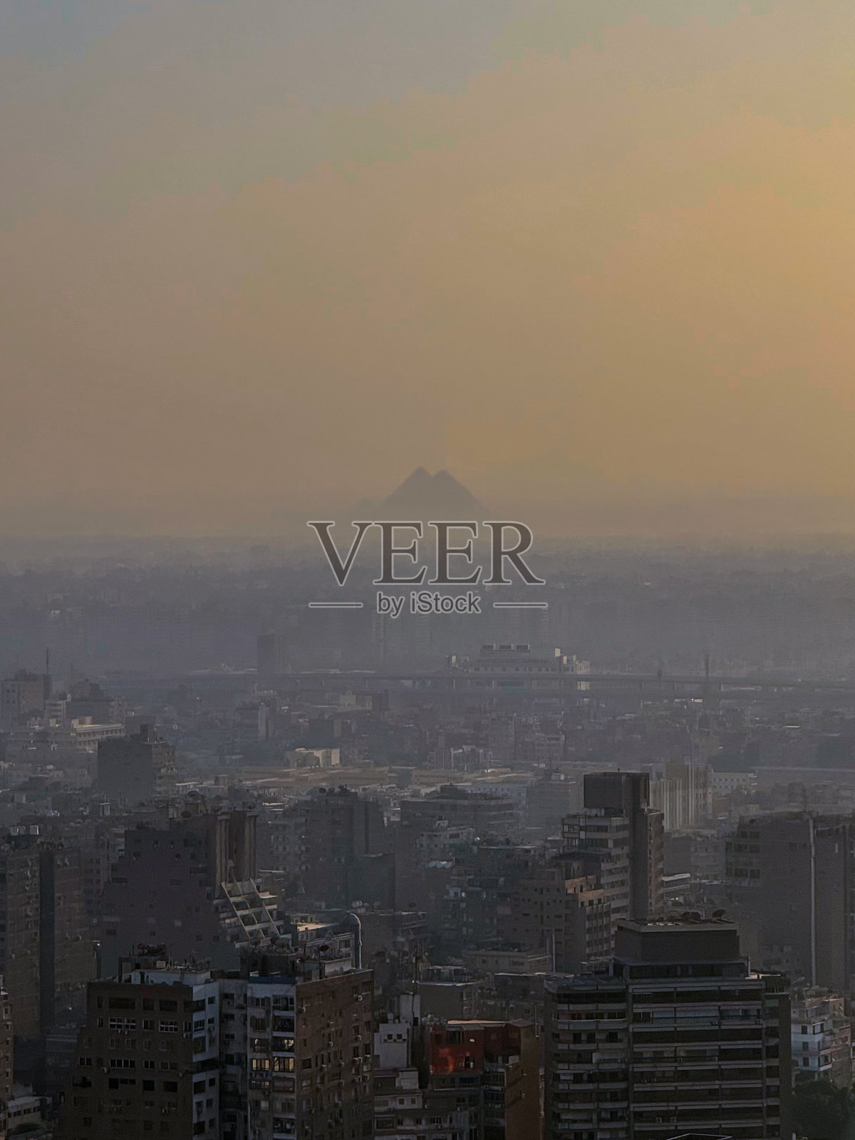 从空中远距离拍摄,吉萨金字塔的剪影在浓重的烟雾中显现,其上方是密集的城市发展。照片摄影图片