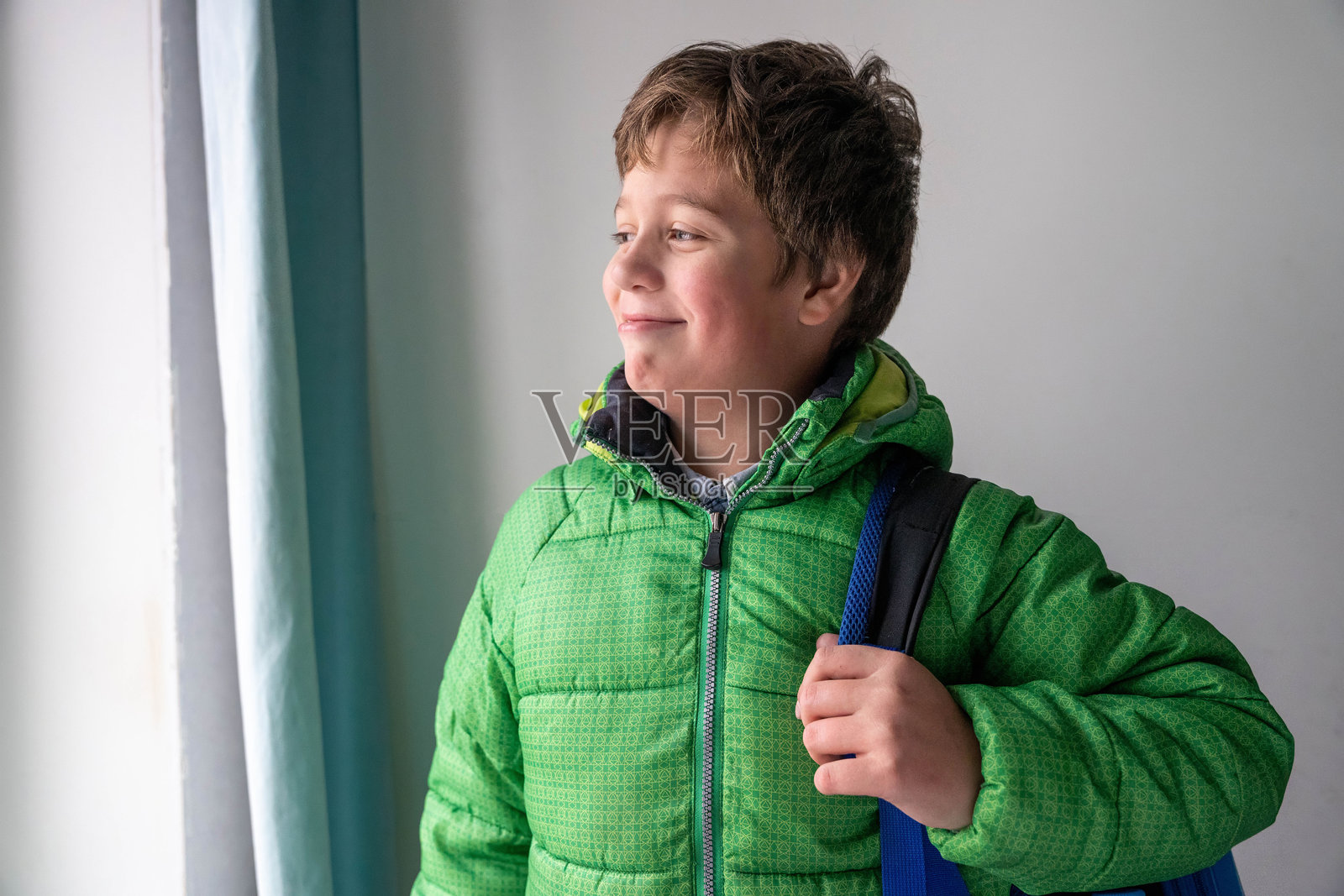 一个穿着绿色夹克的男孩站在窗边,手里拿着包,面带微笑照片摄影图片