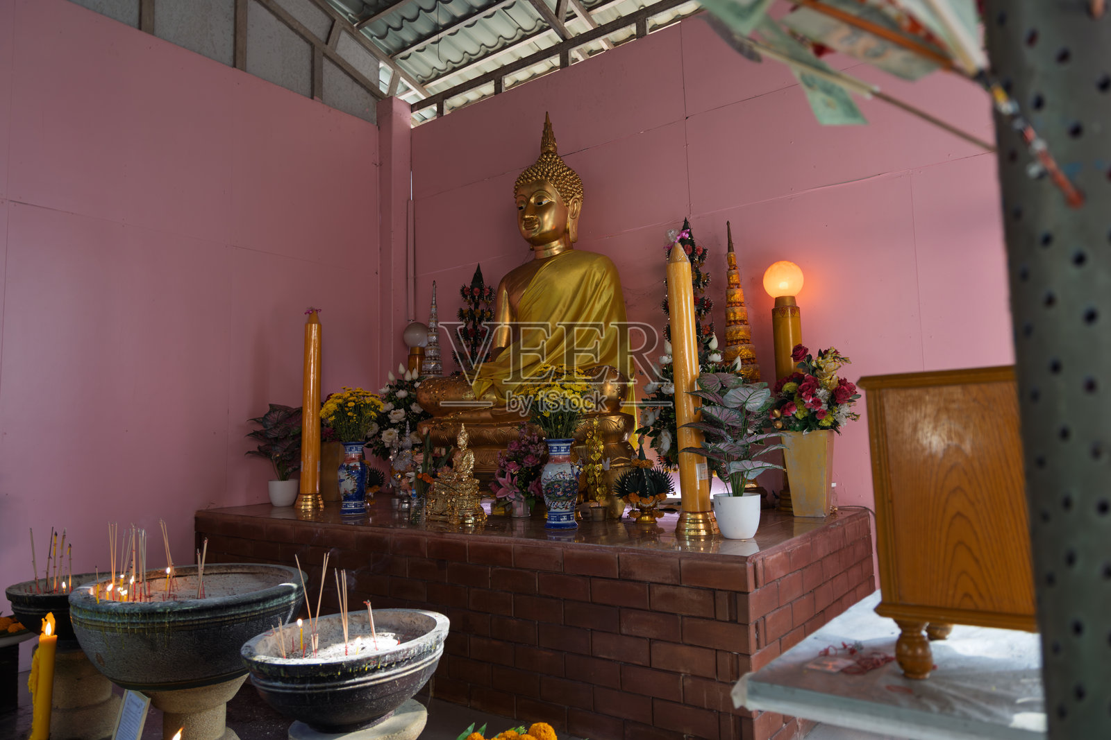 泰国佛教圣地金色佛像,周围环绕着蜡烛、香炉、鲜花供品和微型佛塔,粉色墙壁为背景照片摄影图片