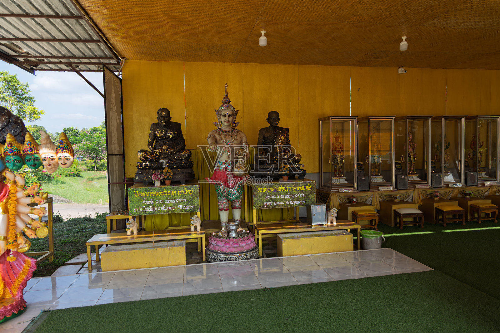 泰国寺庙内三尊坐佛雕像和华丽的守护神像,背景为黄色,摆放着祭祀用品,陈列柜中展示着宗教物品,为户外场景照片摄影图片