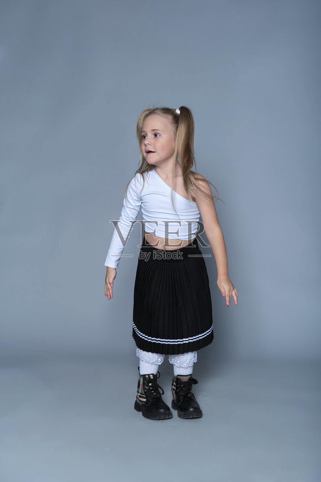 一位金色双马尾的小女孩,身穿白色露肩上衣和黑色百褶裙,自信地站着。她时髦的装扮与天真的神态之间充满趣味的对比,突显了青春时尚。照片摄影图片