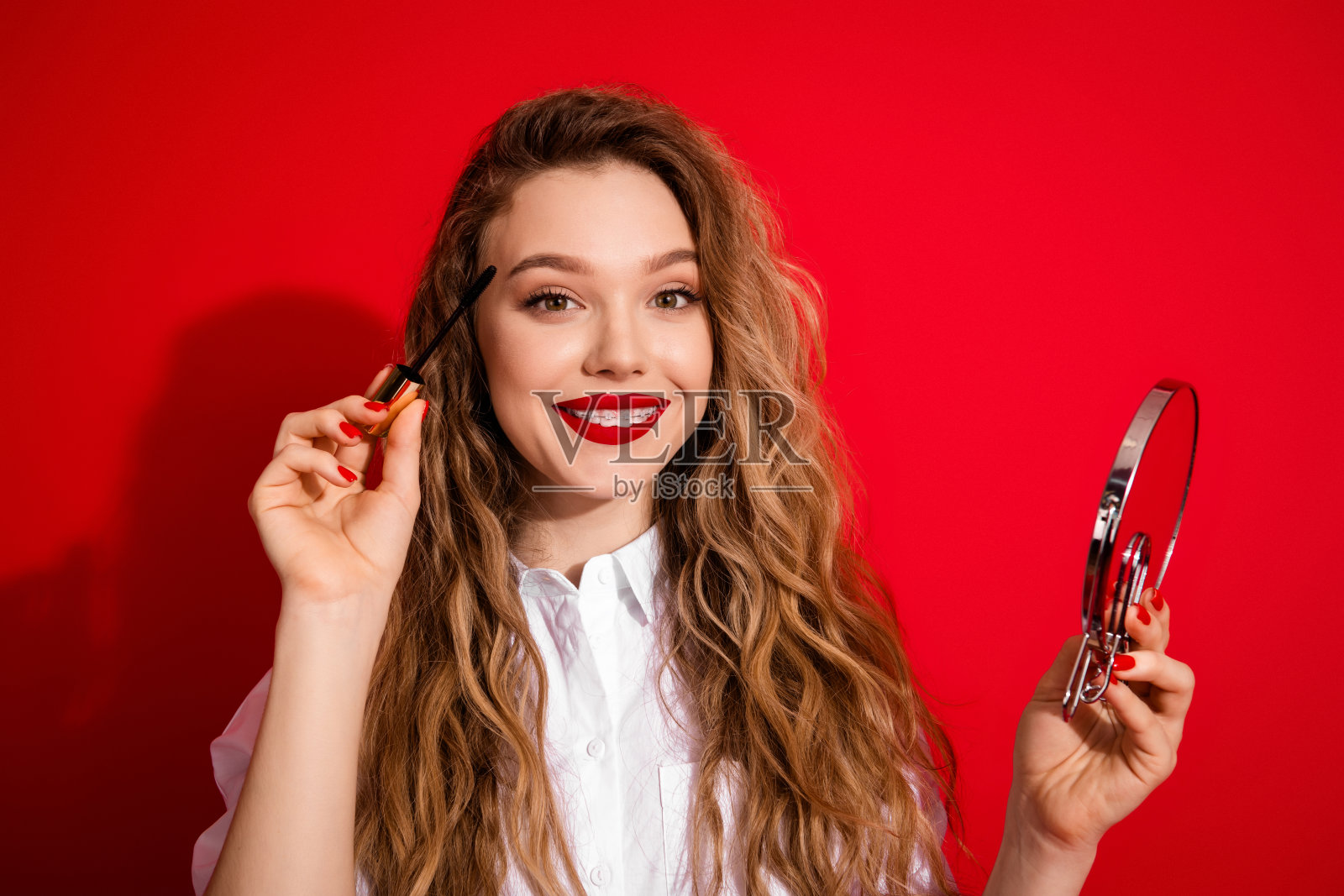 身着白衬衫的魅力女性,在红色背景前,手持睫毛膏刷和镜子化妆照片摄影图片