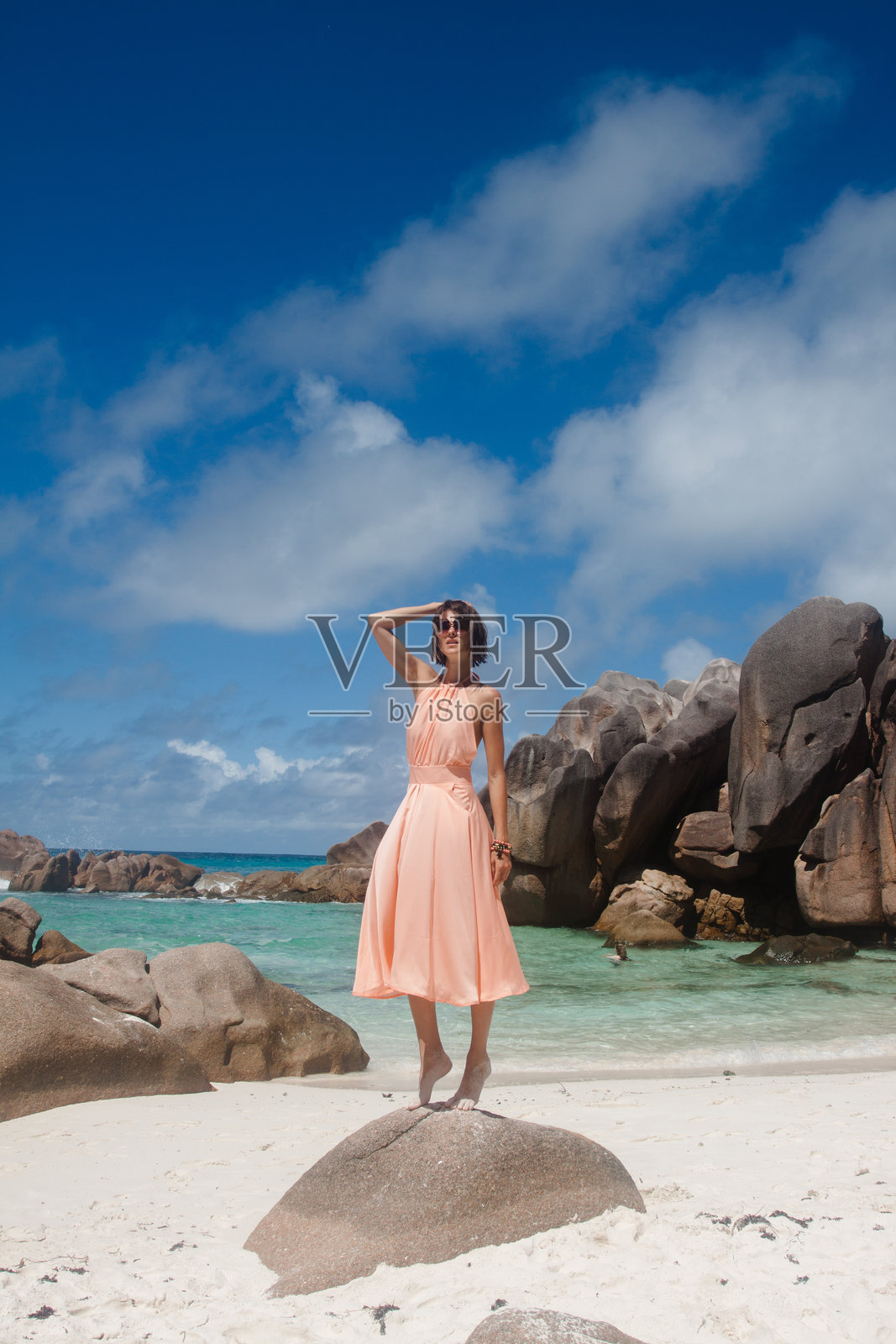 一位身着浅玫瑰粉色浪漫连衣裙的年轻女子,站在白色沙滩边缘的岩石上。她身后是岩石构造、清澈的绿松石般的海水。照片摄影图片