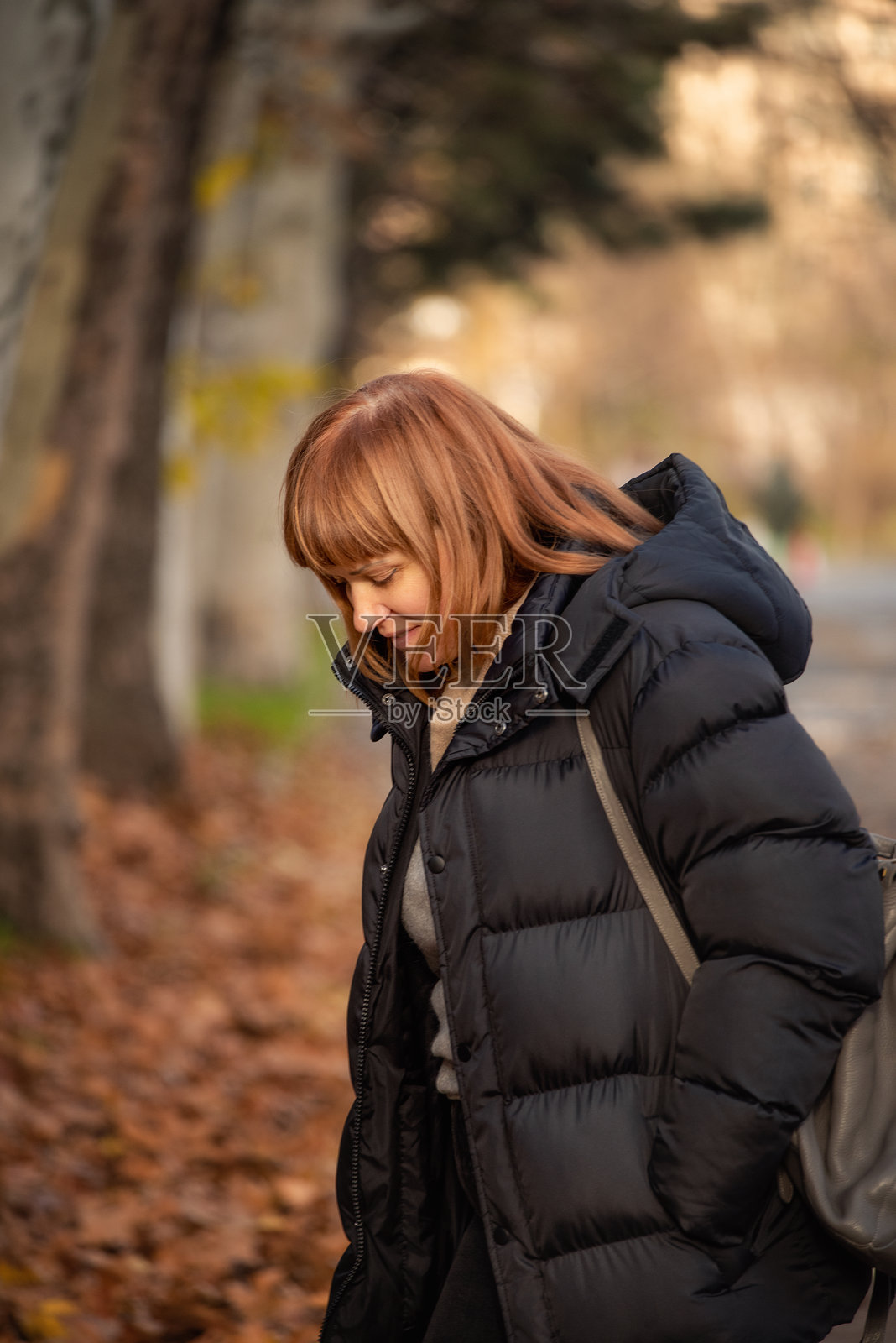 一位姜黄色头发的女士穿着温暖的黑色羽绒服走在秋季公园里。柔和的金色光线、落叶和宁静的日常生活氛围。照片摄影图片