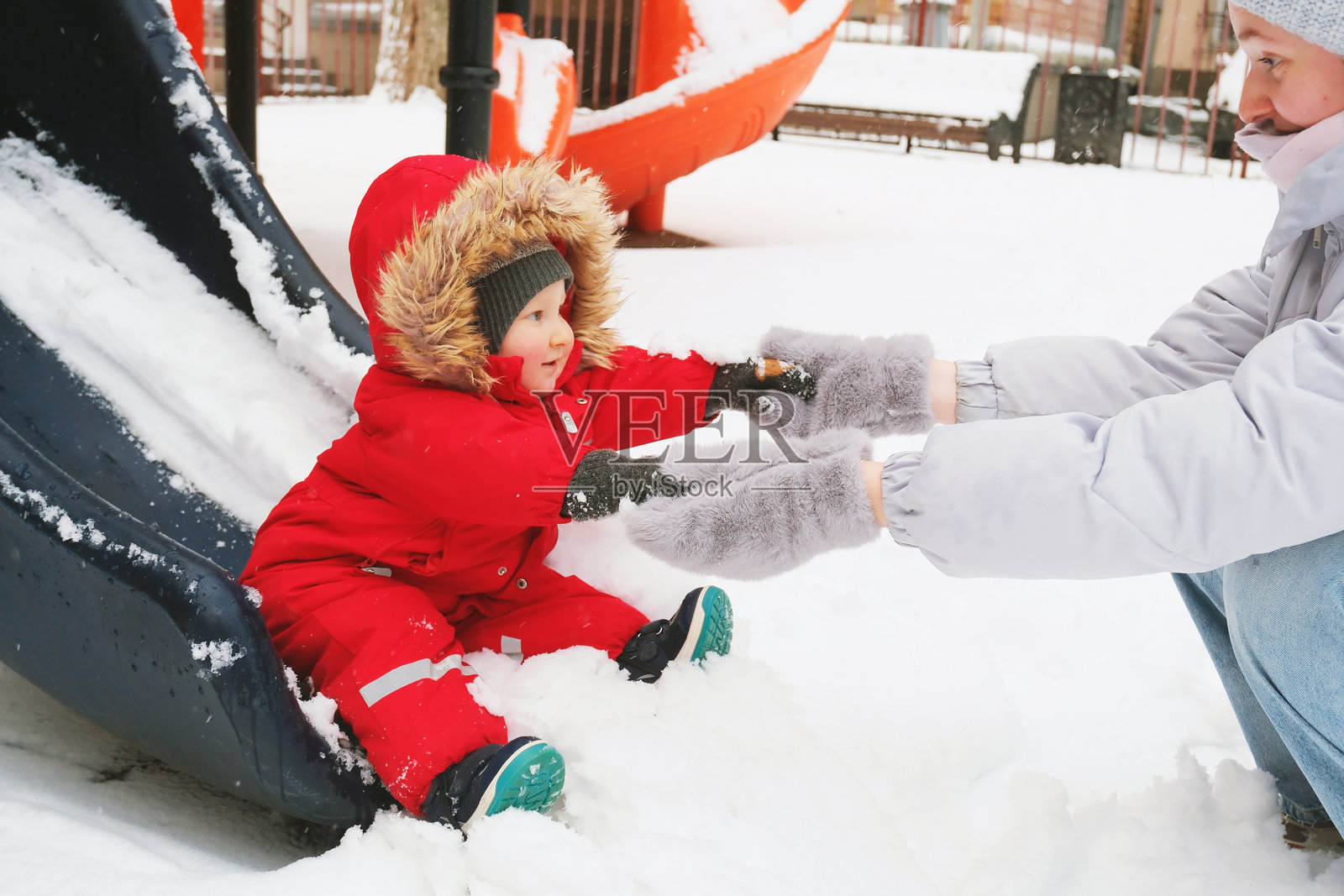 一个穿着红色雪服的孩子在冬季游乐场的雪滑梯上,由一位成年人帮助着。孩子们的笑容和笑声捕捉到了欢乐的互动和季节的喜悦。照片摄影图片