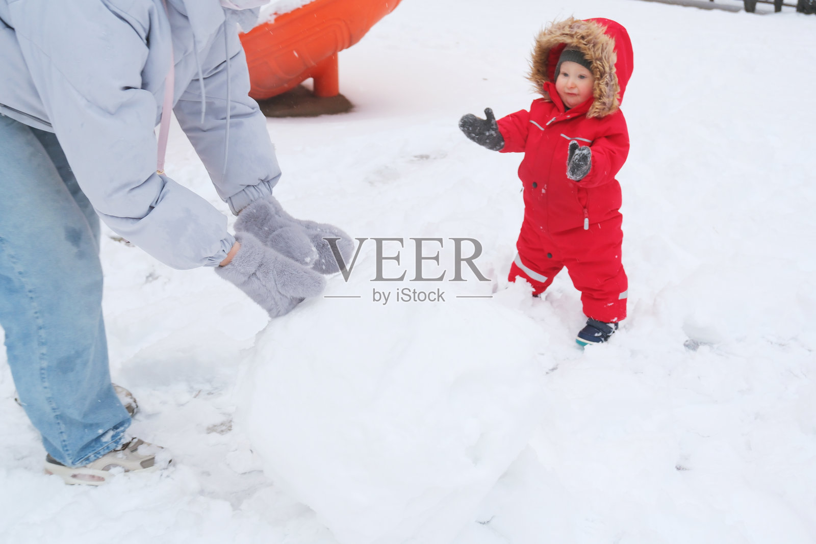 穿着红色雪服的孩童,看着一名成年人在雪地公园里堆雪球。孩童对这有趣的活动显得很兴奋,营造出欢乐的冬日时光。照片摄影图片